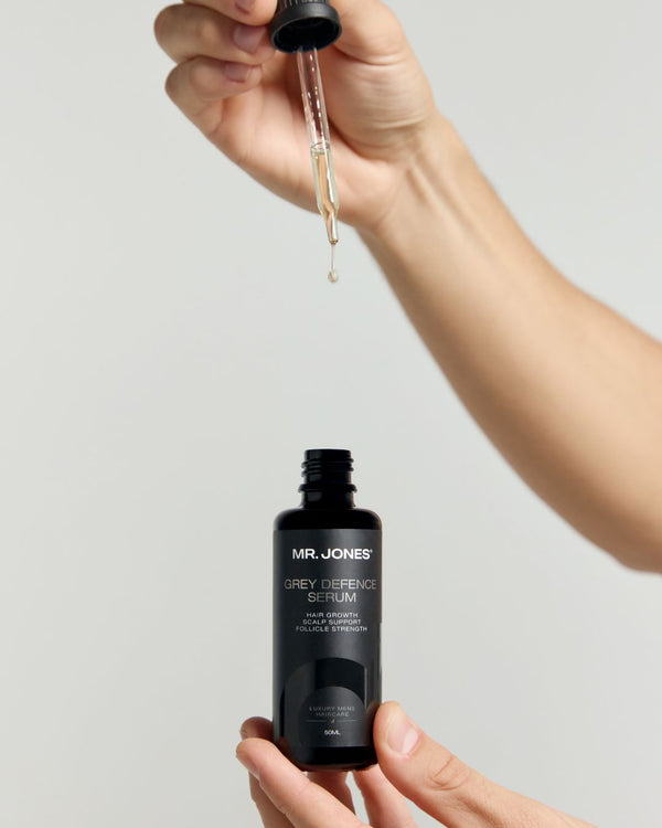 MR. JONES Grey Defence Serum bottle with dropper held by a hand against a light grey background