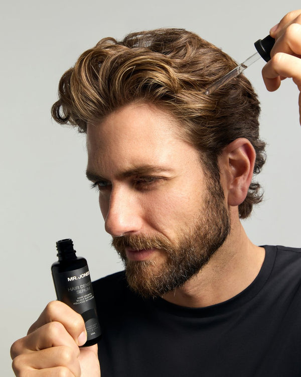 Man applying hair product 'Hair Density Serum' to his hair with a dropper bottle against a neutral background