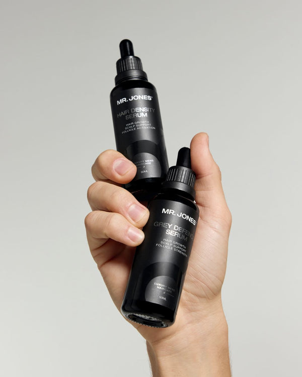 Hand holding two black bottles of Mr. Jones hair density serum and grey defence serum against a grey background