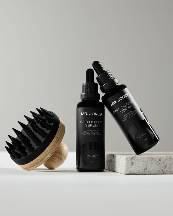 Two black bottles of 'MR. JONES' hair density serum and Grey Defence Serum with a brush on a light grey background