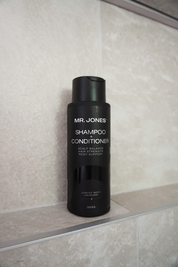 A black bottle of Mr. Jones Shampoo + Conditioner 2-in-1 standing on a light beige tiled shelf.