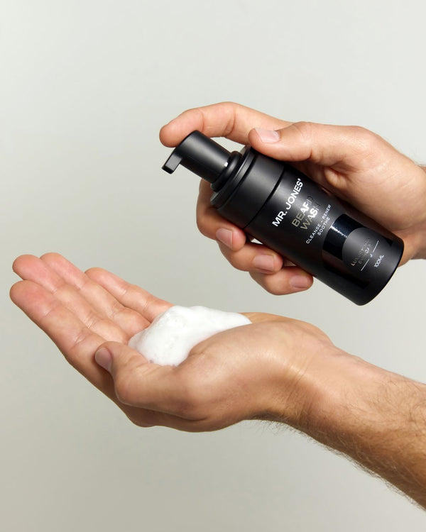 Hand holding a black bottle labeled 'Mr. Jones Beard Wash' with foam on a light gray background